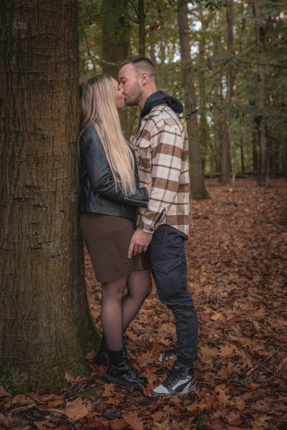 A couple shares an intimate moment against a tree trunk in an autumn forest with fallen leaves on the ground.