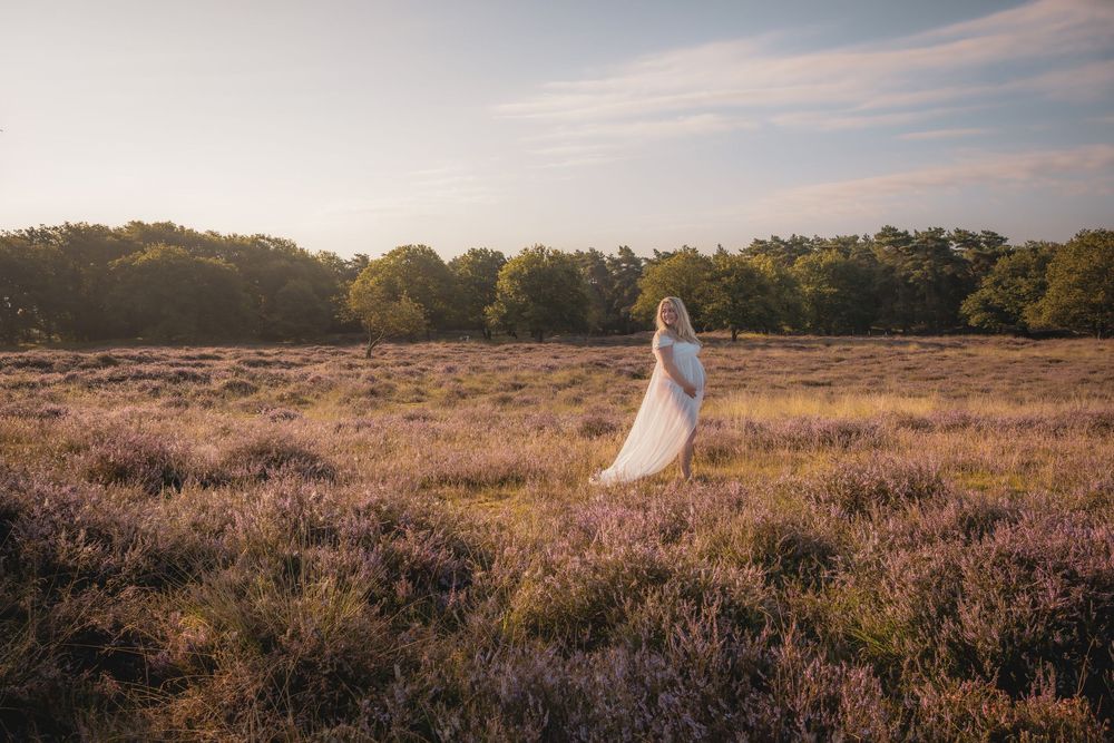 A figure in a flowing white dress stands in a golden field at sunset with trees in the background.