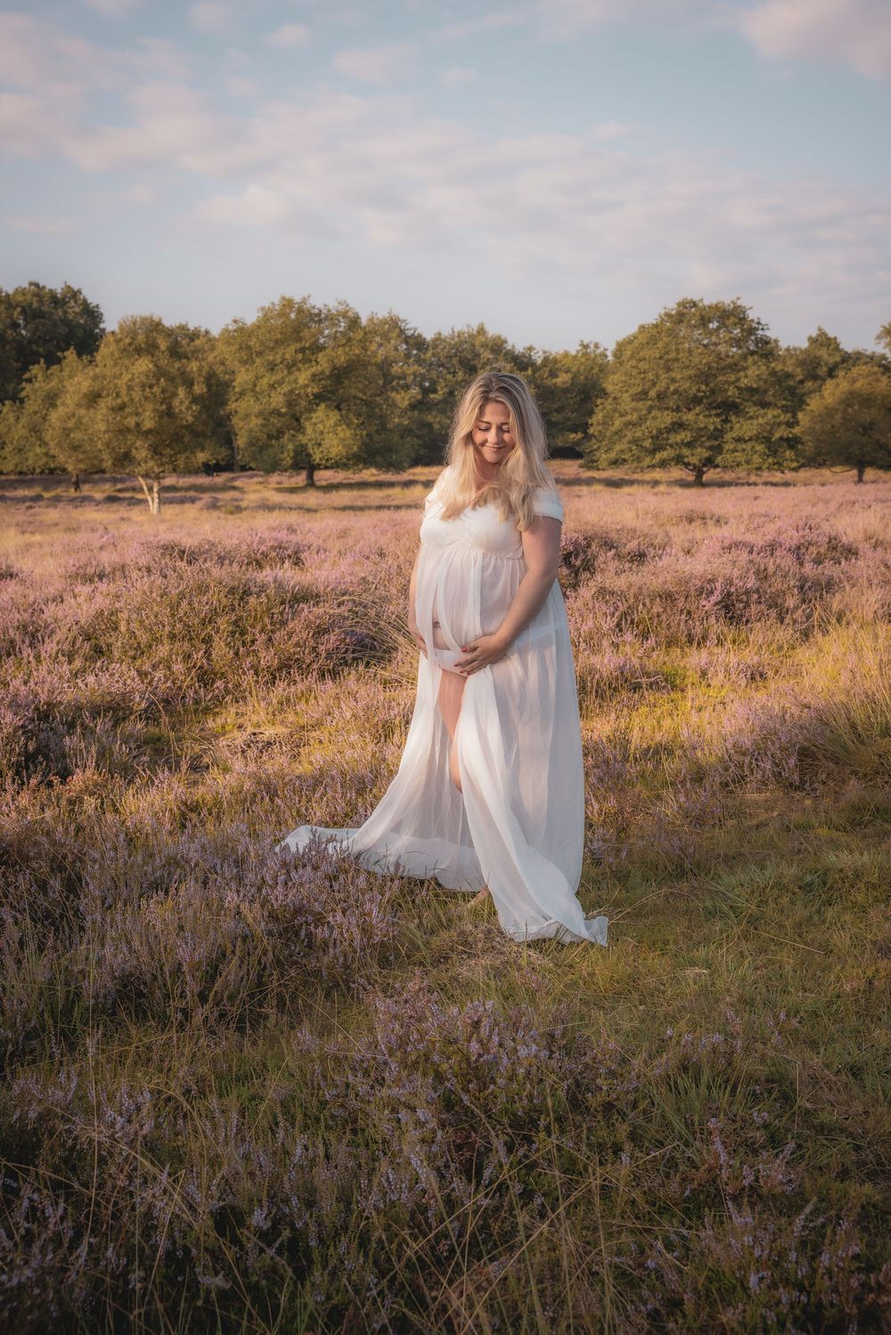 A figure in a flowing white dress stands gracefully among purple heather fields during golden hour.