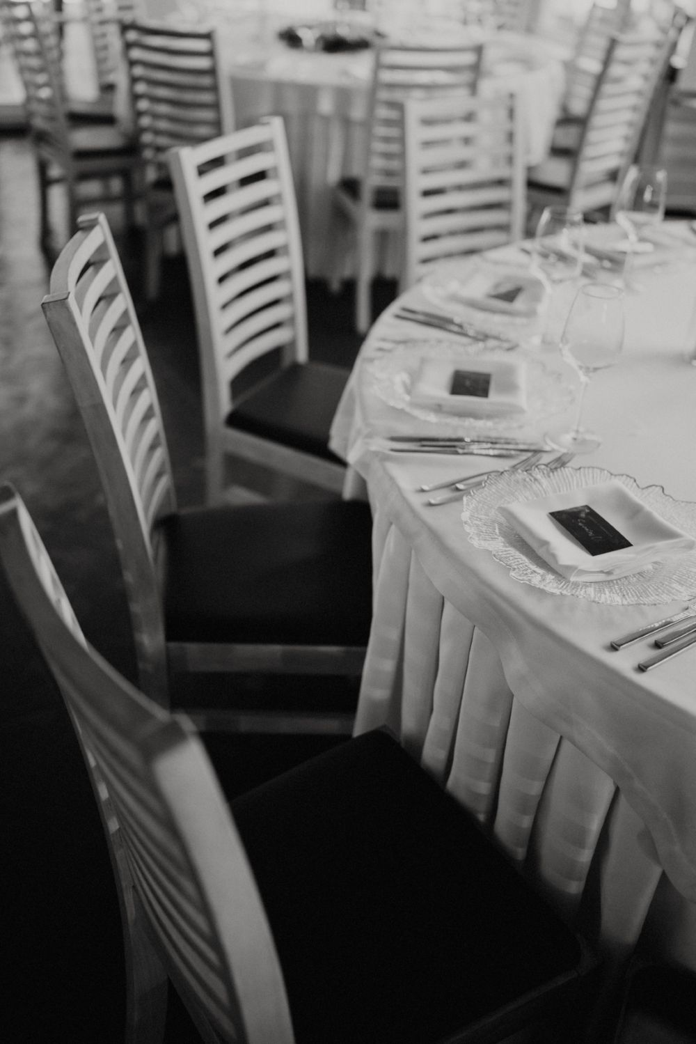Black and white photo of formal table settings with white linens and ladder-back chairs.