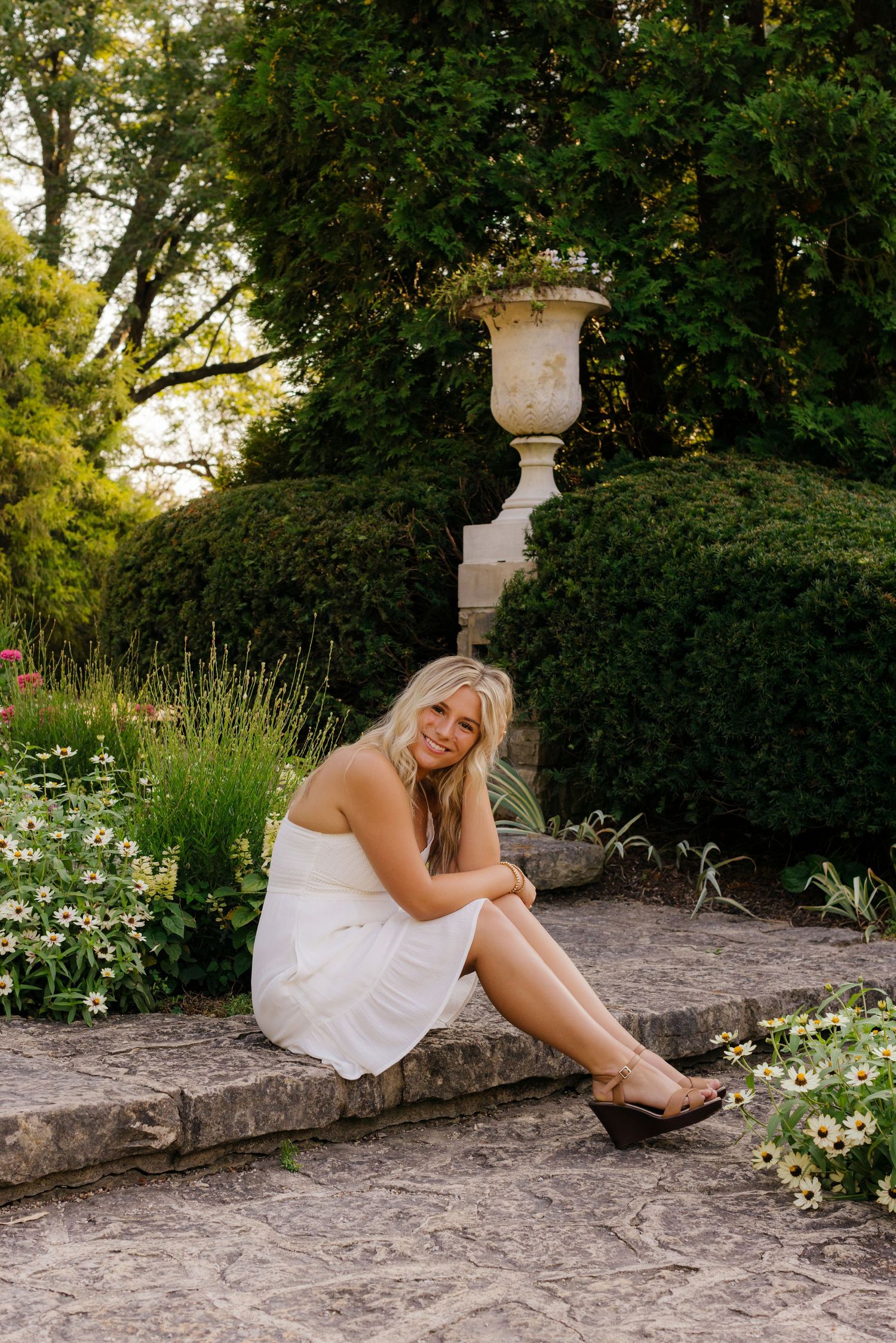 Woman in white dress sits on stone ledge in elegant garden with decorative urn and blooming flowers.