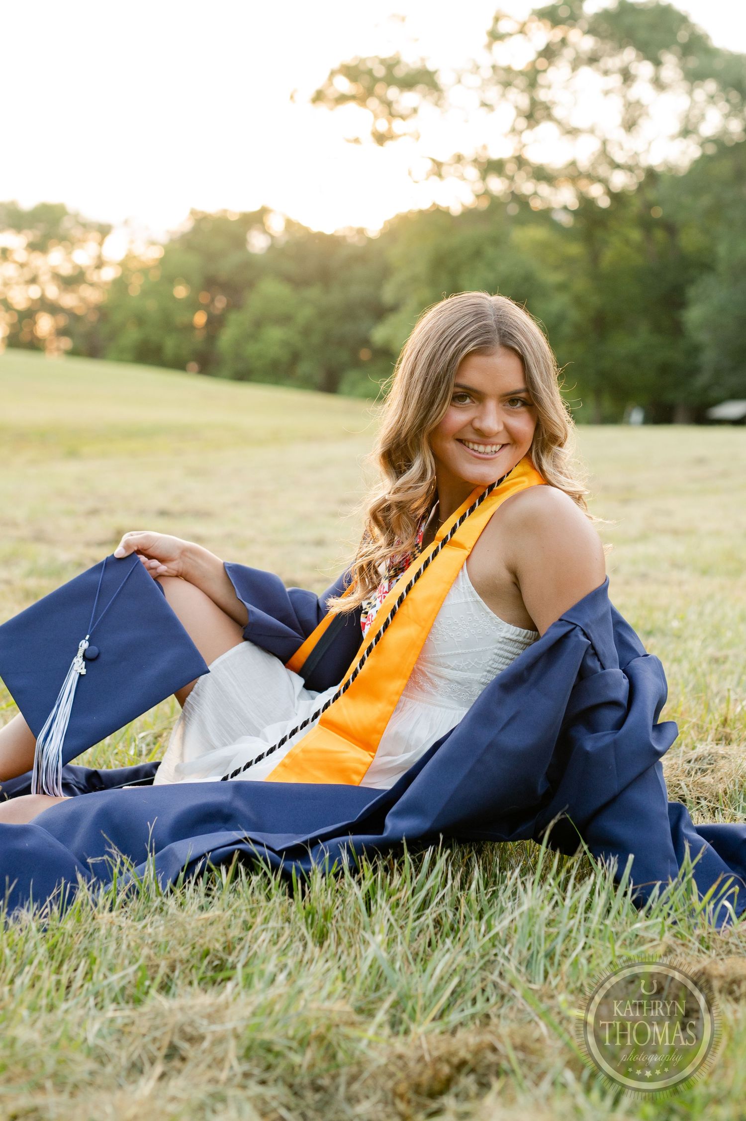 A graduate lounges in grass wearing a yellow stole and navy graduation gown at sunset.