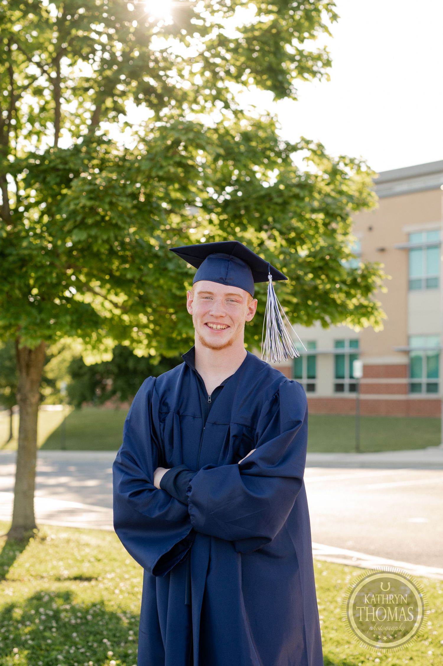 Graduate in academic regalia poses outdoors with sunlit trees and building in background.