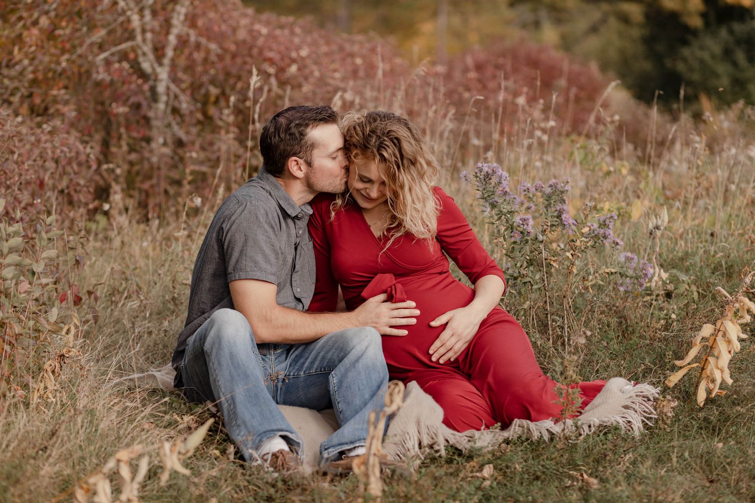 Couple sits in tall, fall colored grasses. He touches her belly and kisses her on the cheek at Cataraqui Creek in Kingston, Ontario.
