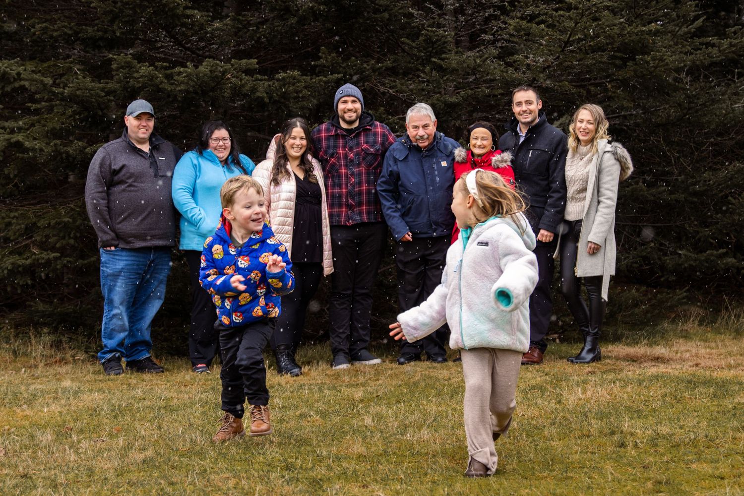 An extended family gathers outdoors by evergreen trees while two children in winter coats play in the foreground.