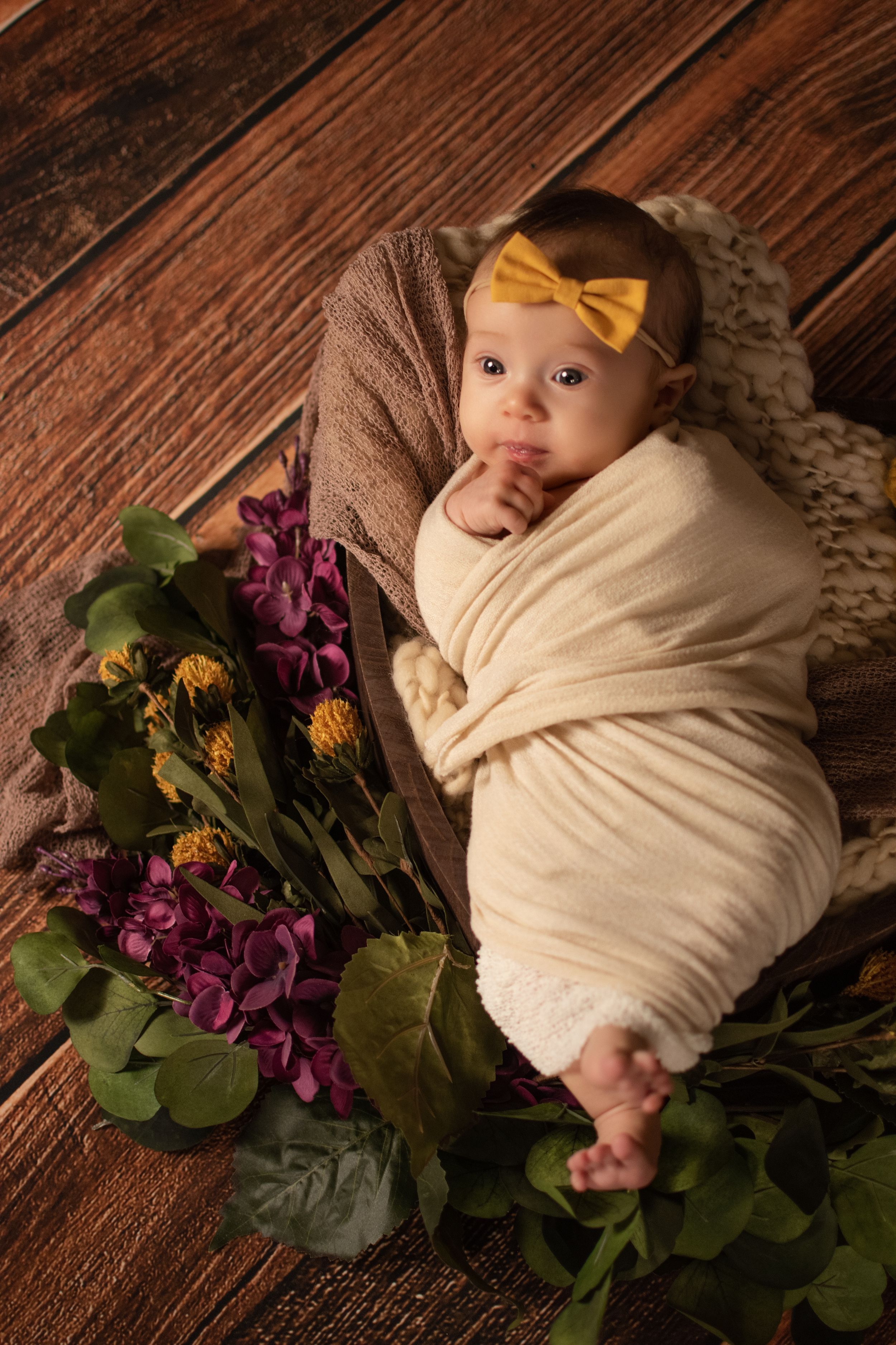 Newborn wrapped in cream fabric with yellow bow accent and purple flowers on wooden surface.