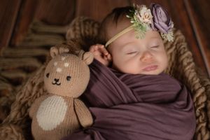 Cozy newborn portrait with plush bear toy and purple wrap on textured background.