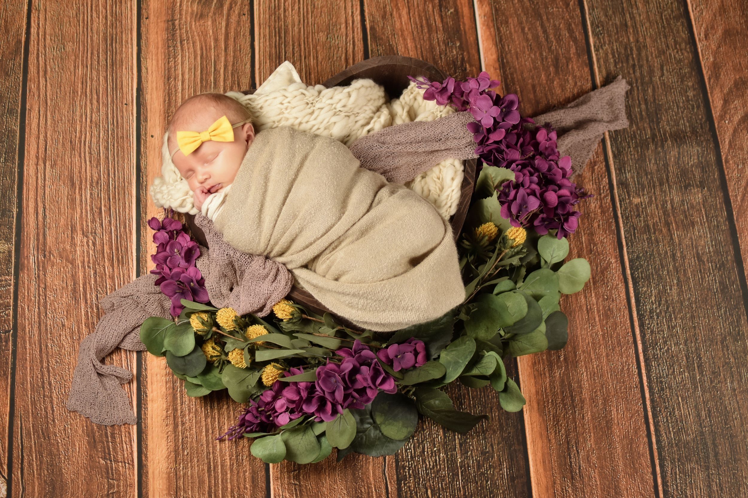 Newborn baby wrapped in beige blanket lying on wooden surface with purple flowers and greenery.