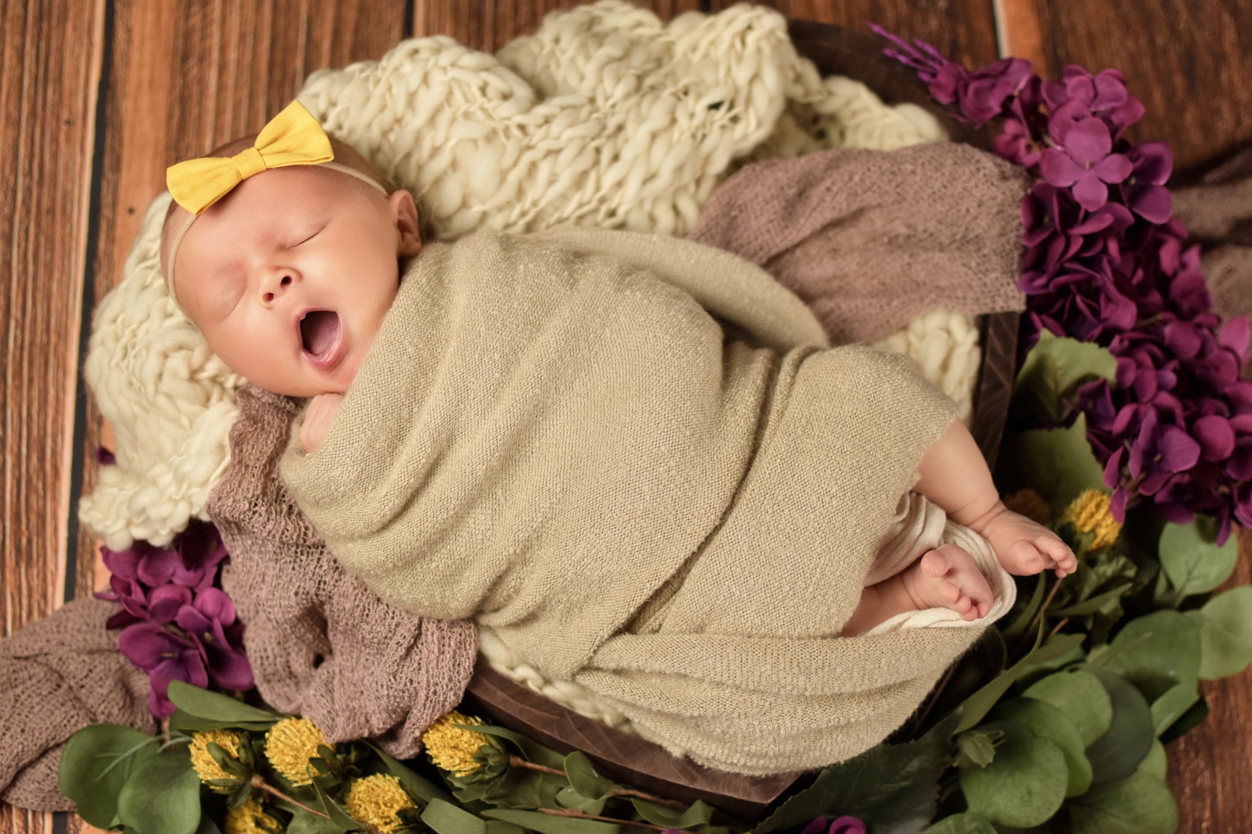 Sleeping newborn in cream wrap with yellow bow and purple floral decorations on wooden background.