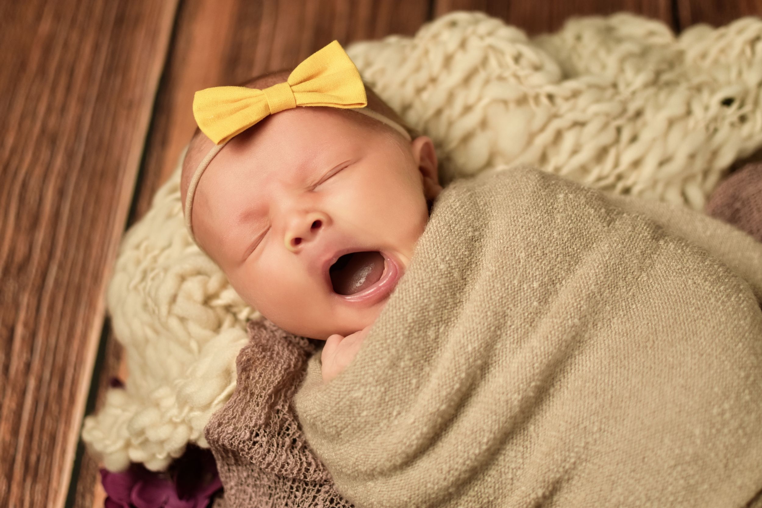 Side view of sleeping newborn with yellow bow accessory on wooden surface with purple flowers.
