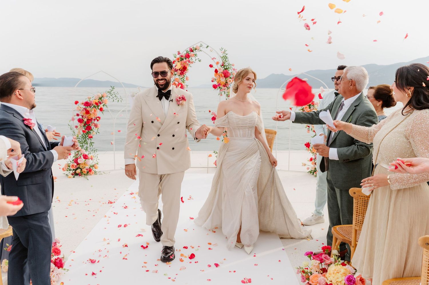A wedding ceremony takes place under a rustic floral arch on a hillside with guests seated on both sides of the aisle.