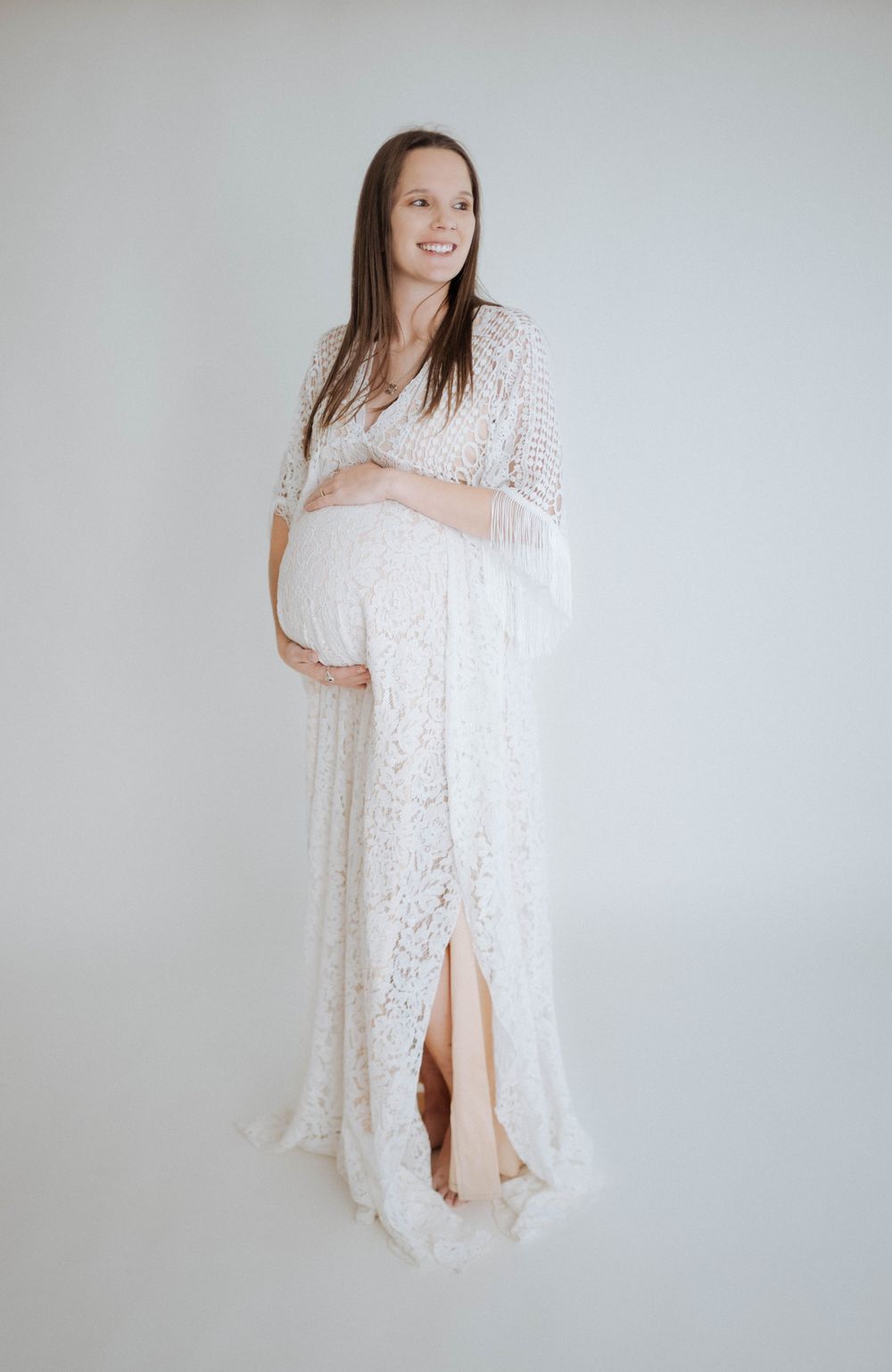 A pregnant person in a white lace maternity dress poses against a light gray background for a studio portrait.