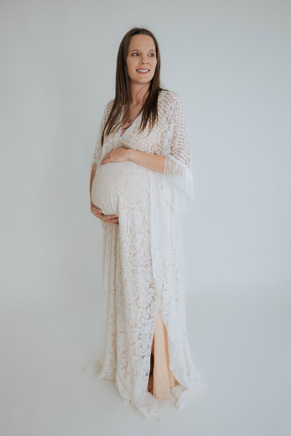 Expectant mother wearing flowing white lace maternity dress poses against light grey background in studio setting.