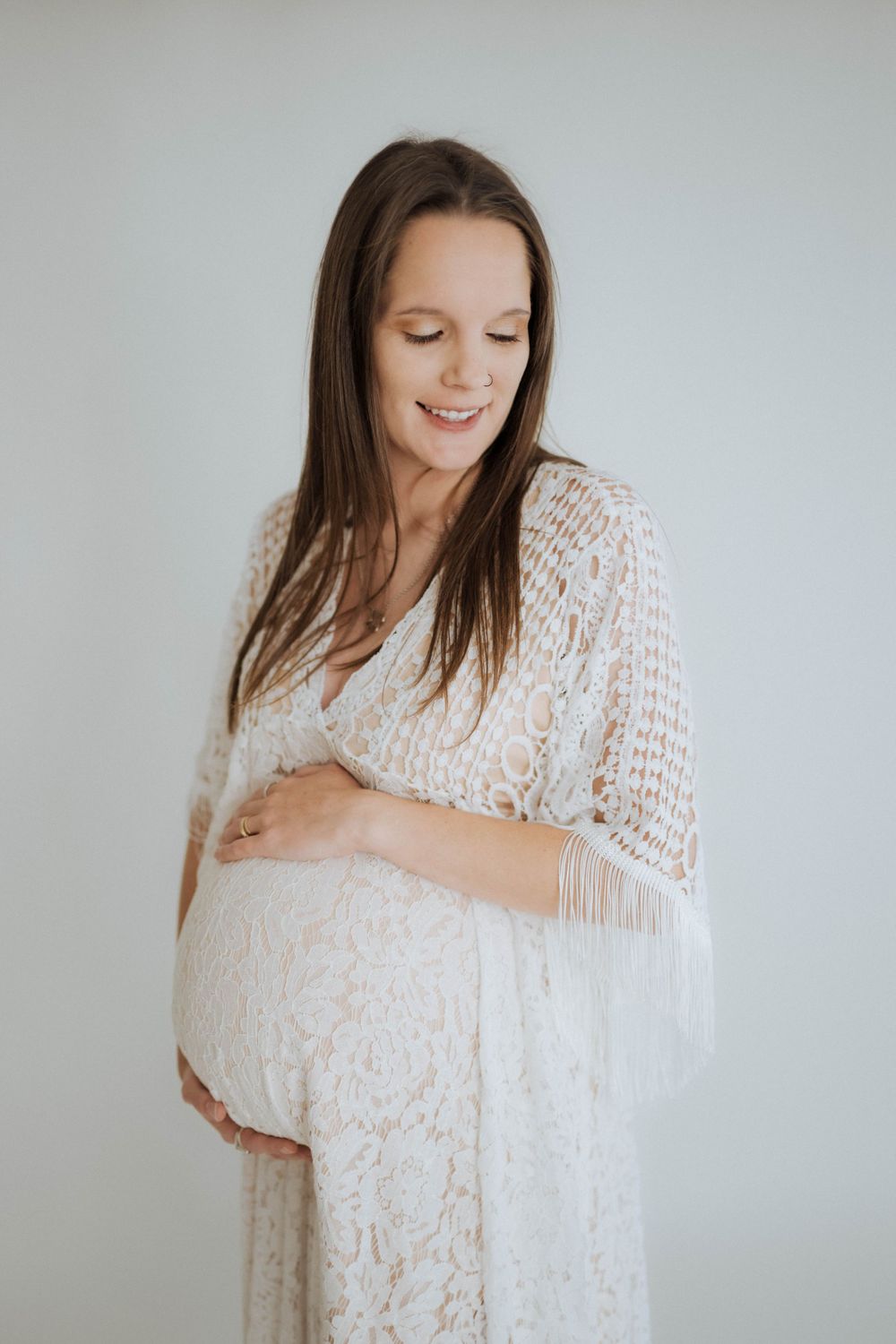 A pregnant woman in a white lace dress gently touches her belly while smiling with eyes closed.