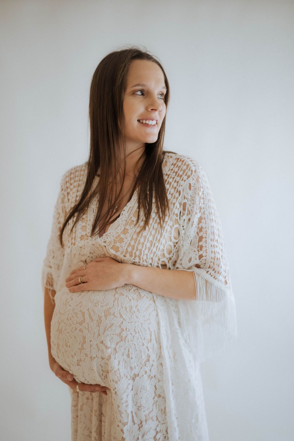Expectant person in a delicate white lace dress poses against light background during maternity photoshoot.