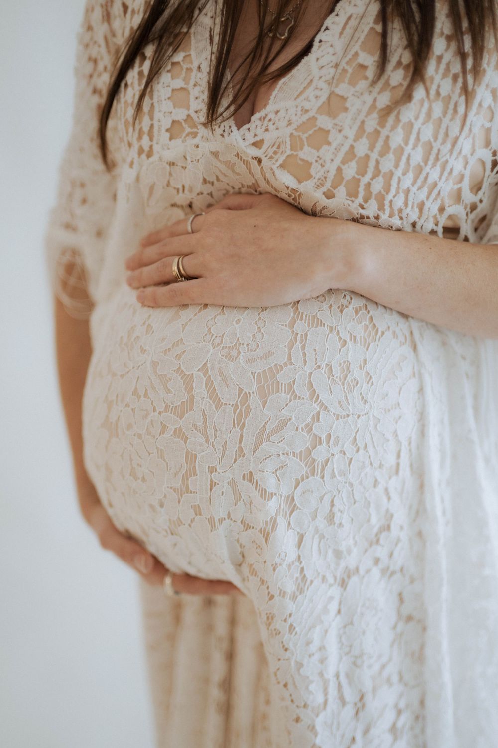 Close-up of pregnant woman's belly in white lace dress with hand resting on bump, wearing engagement ring.