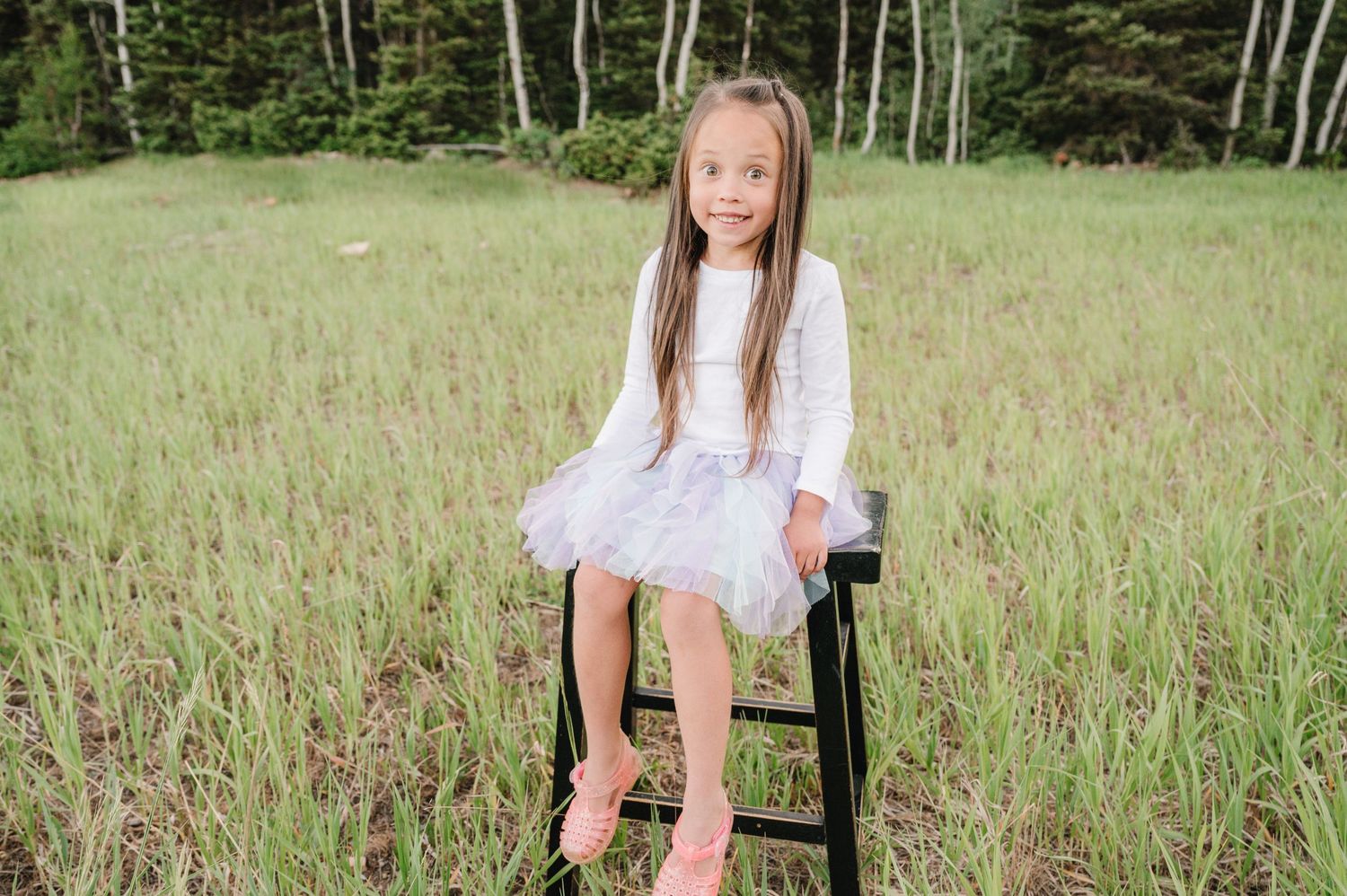 Young girl in white dress sits on black stool in summer meadow with aspen trees in background.