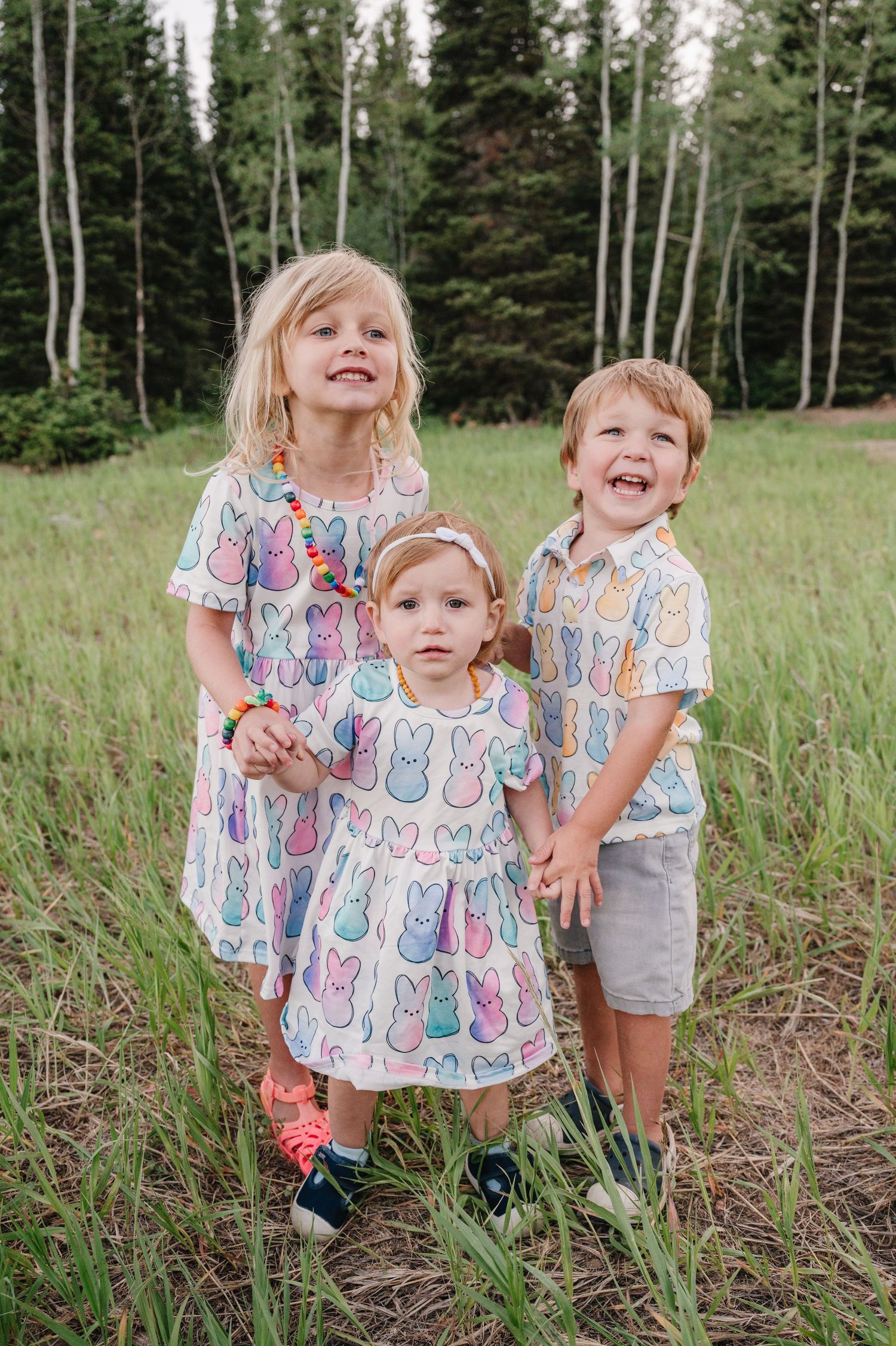 Three young children in matching bunny print outfits stand together in mountain meadow.