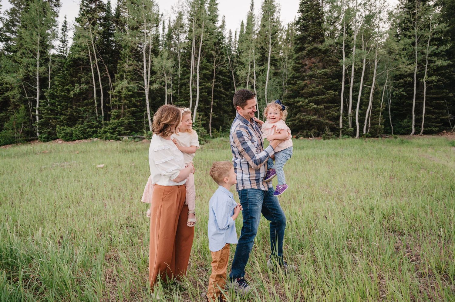 Family of five stands together in mountain meadow with aspen and evergreen trees behind them.
