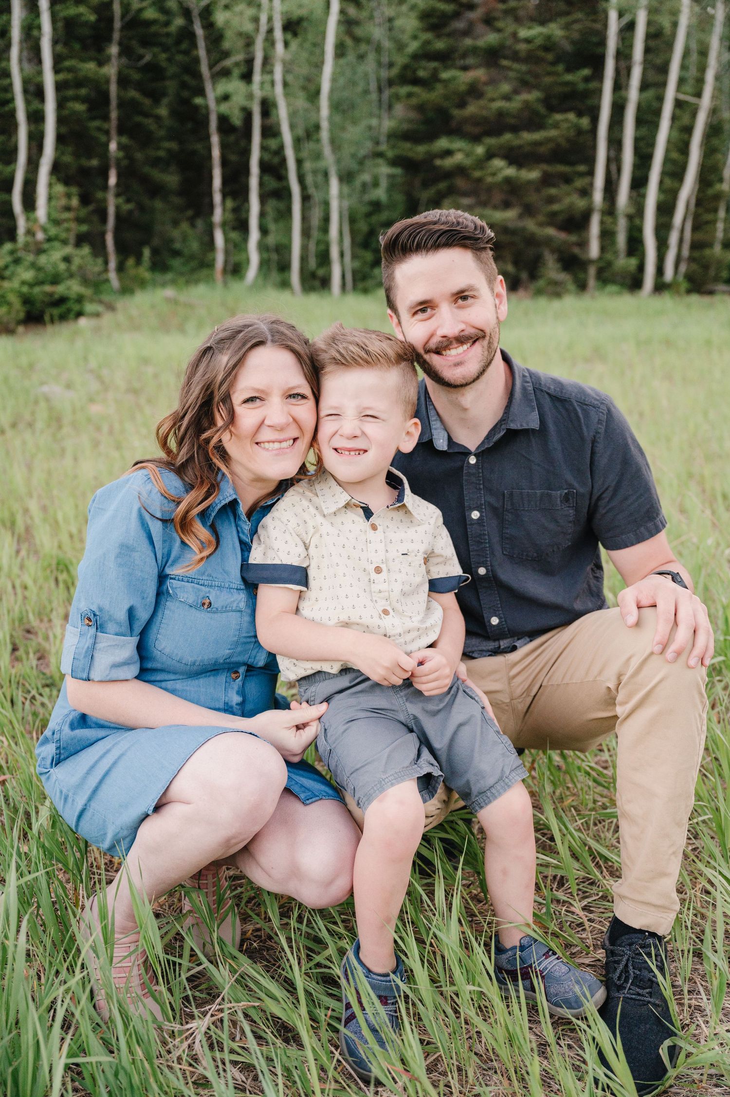 A family of three poses together in a grassy meadow surrounded by aspen trees.