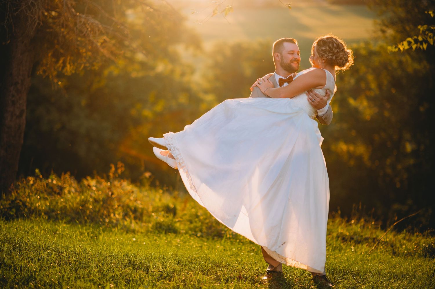 A couple in wedding attire dancing in a golden sunlit meadow at sunset, with the bride's white dress flowing in motion.