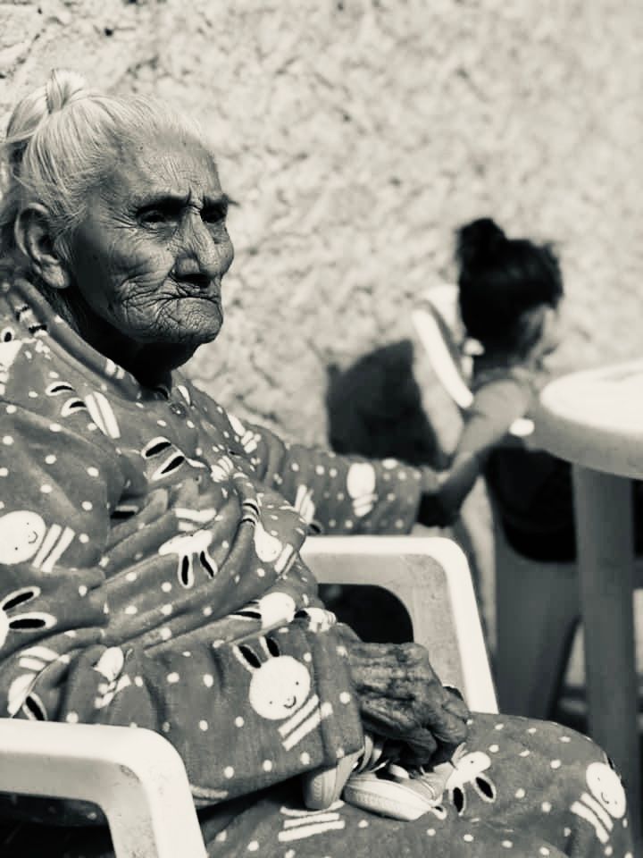 Elderly woman in patterned dress seated outdoors in black and white photograph.