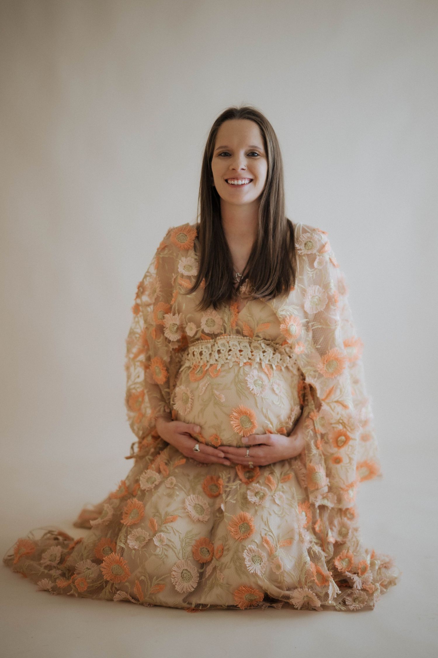 A pregnant woman in a peach-colored floral lace dress poses gracefully against a neutral background in a maternity photo.