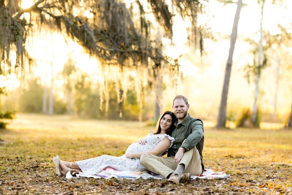 A couple sits on a blanket in a sunlit park with Spanish moss-draped trees during a golden hour maternity photo session.