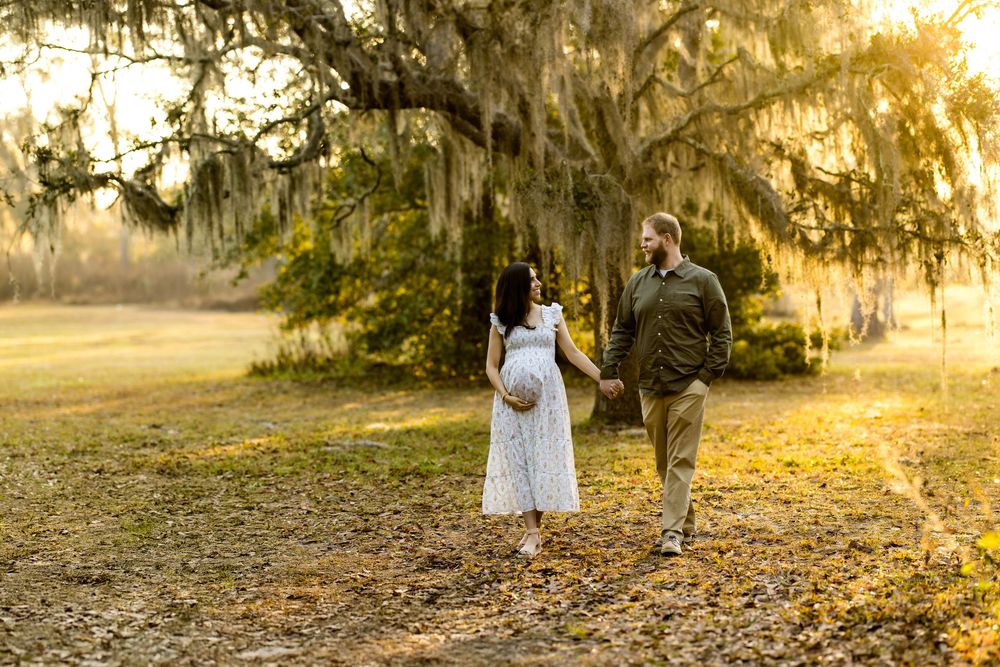 Pregnant couple holds hands walking under Spanish moss-draped trees in golden hour light during outdoor maternity photo session.