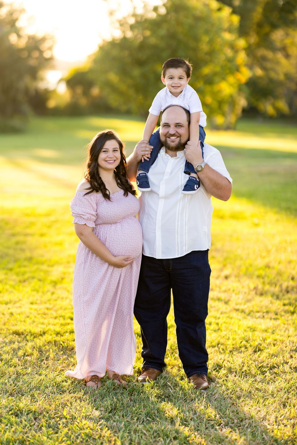A family poses together in a grassy field during golden hour sunset, wearing pink and white outfits.