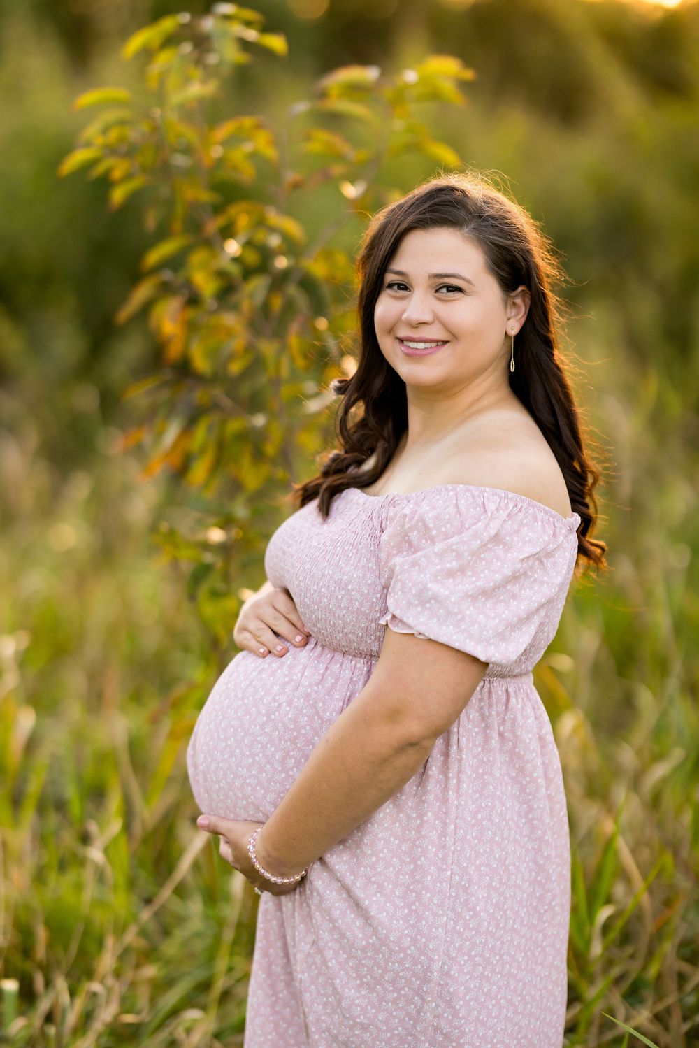 A person wearing a pink polka dot dress poses outdoors in a natural setting with yellow flowers and greenery.