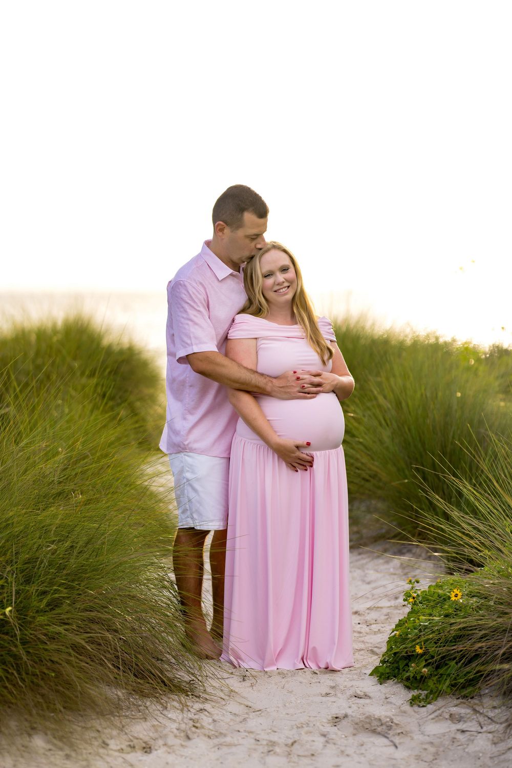 Pregnant couple embracing on sandy beach path surrounded by green dunes, woman in pink maternity gown, man in pink shirt.