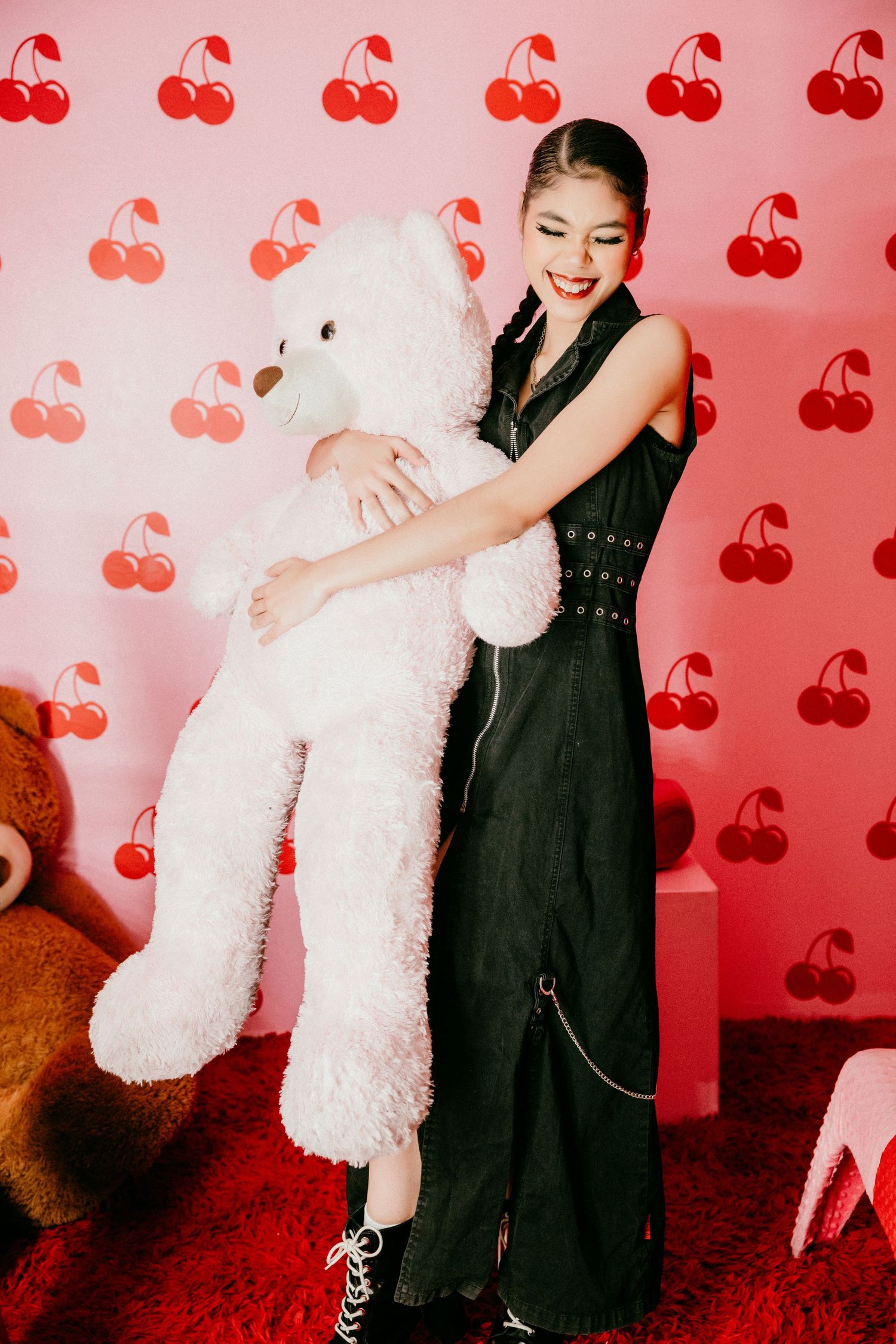 Black jumpsuit-clad person hugging large white teddy bear against pink wall with cherry pattern background.