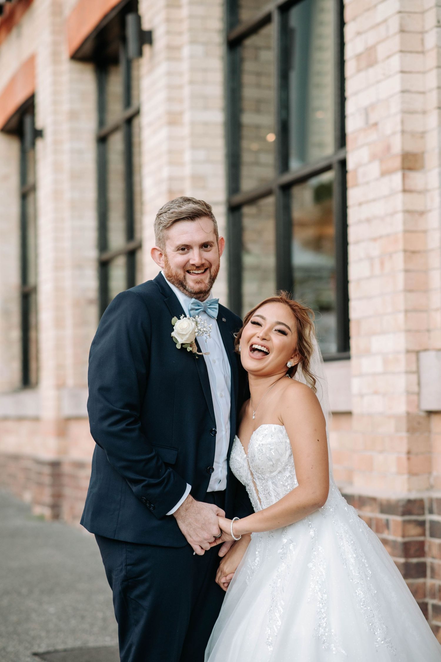 A joyful wedding couple shares a candid moment outside a brick building.