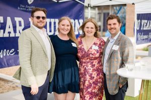 Four people in business attire pose together at a branded event with a blue banner backdrop.