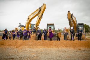 Crowd gathered at groundbreaking ceremony with two yellow excavators on construction site.