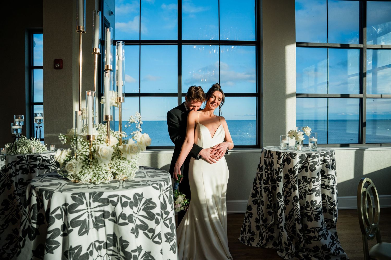 A couple embraces by tall windows in an elegant reception venue with floral patterned tablecloths and ocean views.