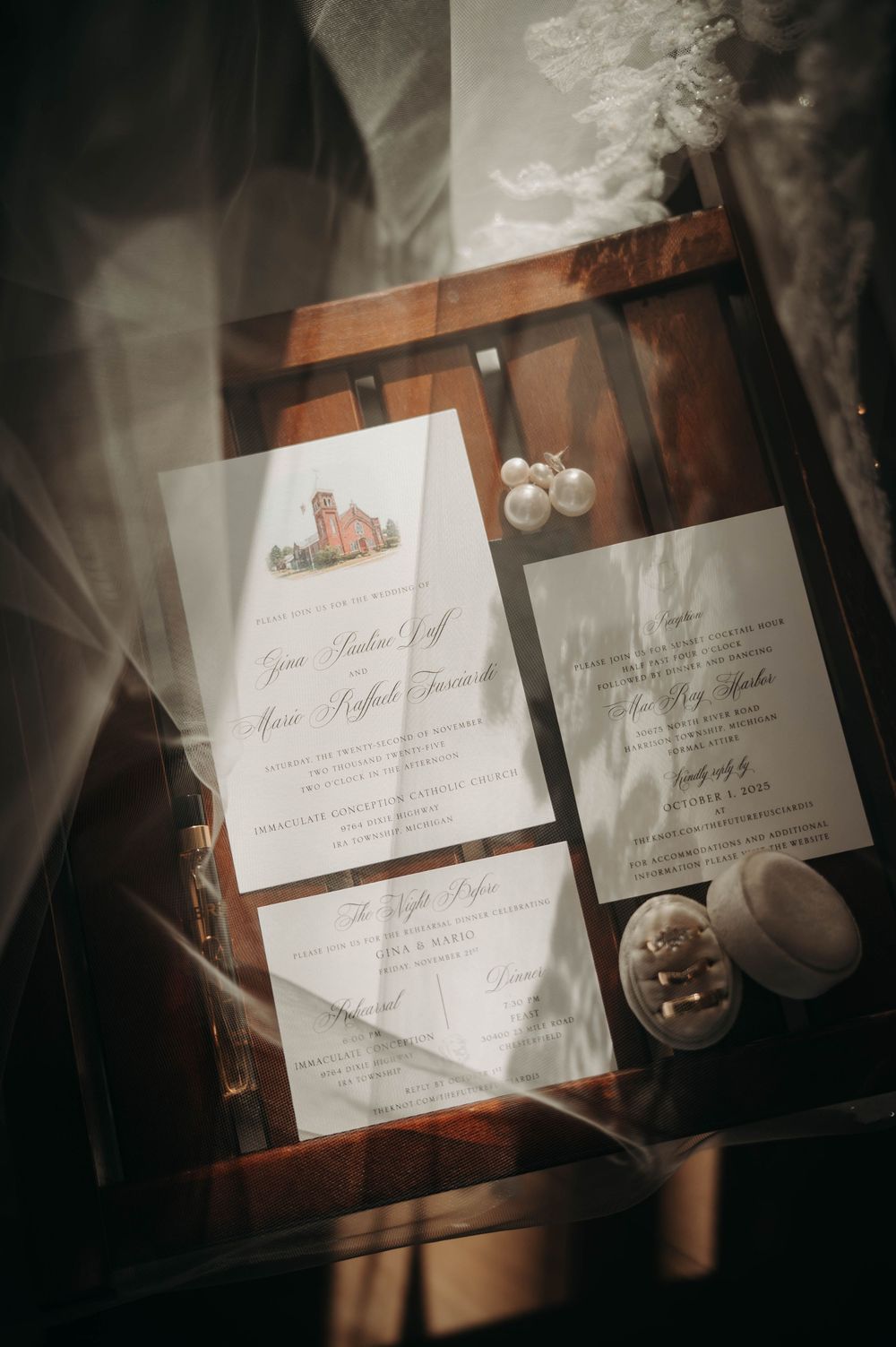 Wedding invitation suite with watercolor church motif, pearl earrings, and ring box on wood chair.