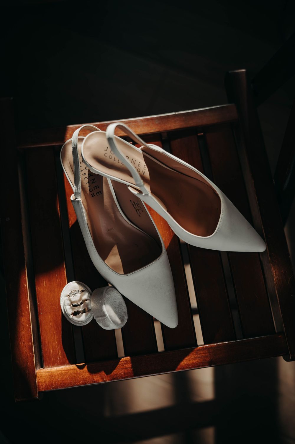 White slingback bridal heels displayed on a wooden chair alongside a decorative ring holder.