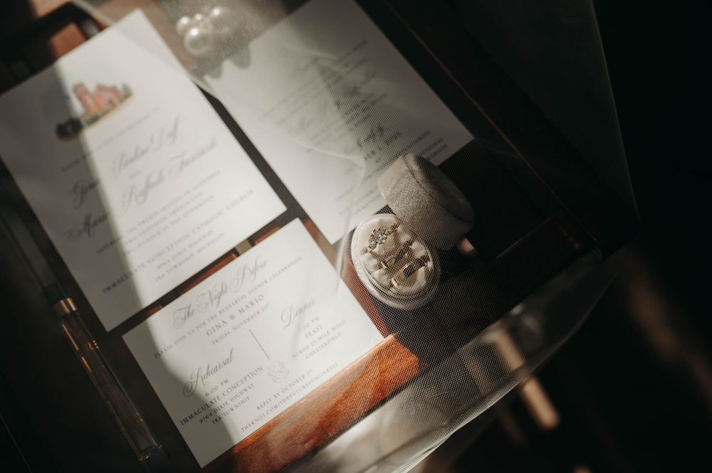 Wedding invitation suite and velvet ring box with diamond rings displayed on a glass surface in sunlight.