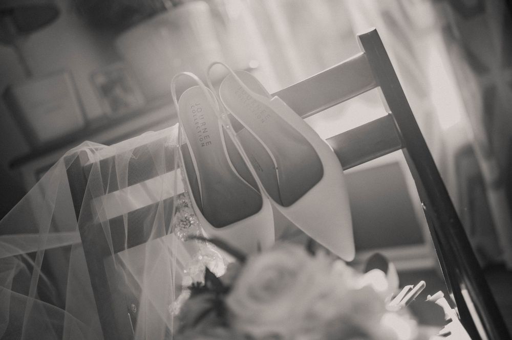 Black and white photo of bridal heels on a chair with veil and white floral bouquet nearby.