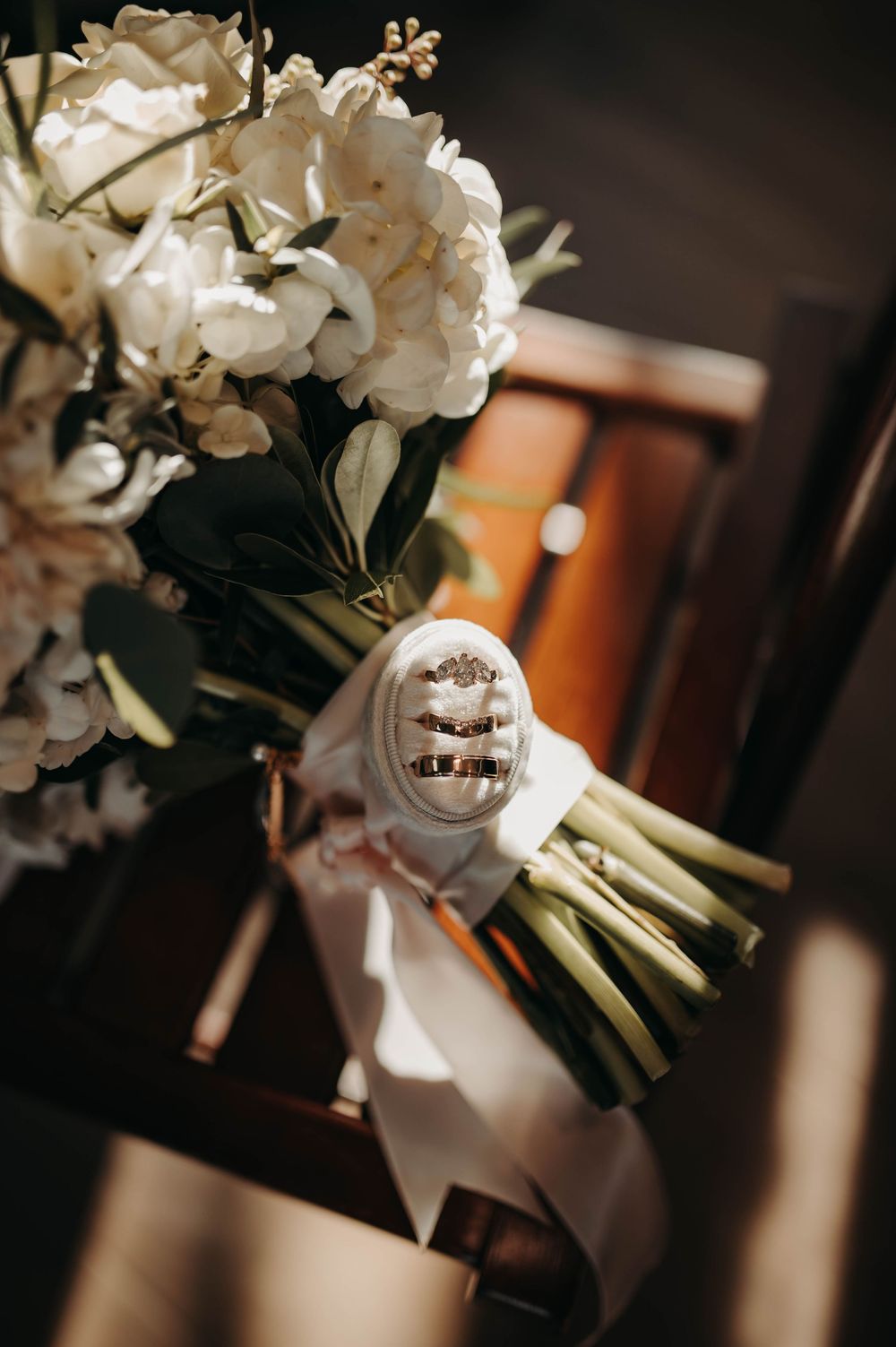White floral bridal bouquet with ribbon and velvet ring box holding wedding bands resting on church pew.