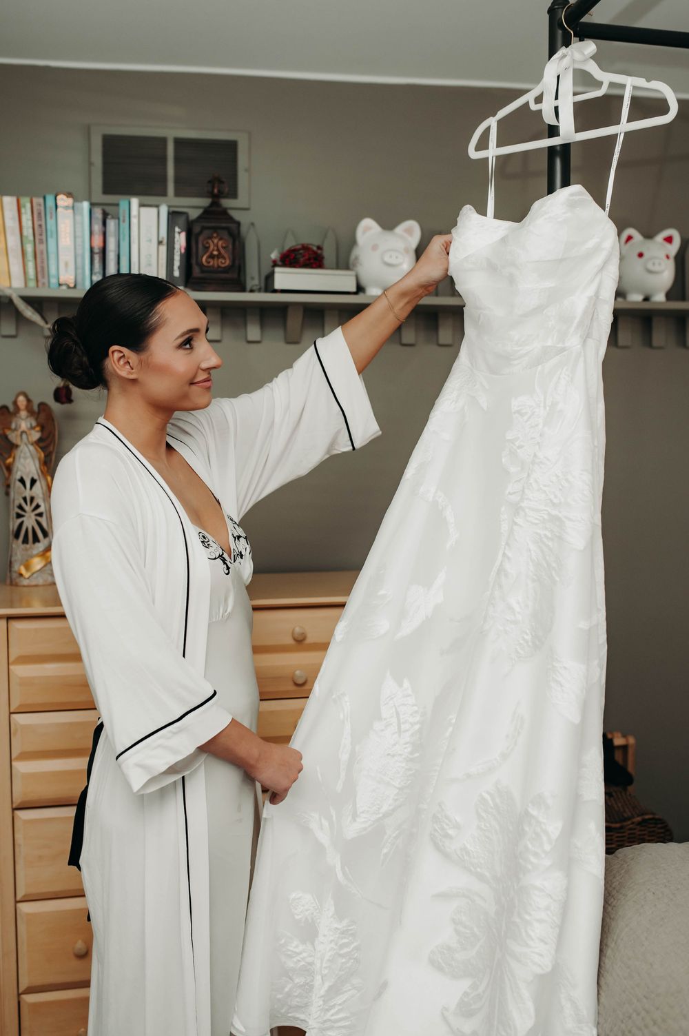 Bride in white robe admiring her strapless lace wedding dress hanging in a cozy bedroom.