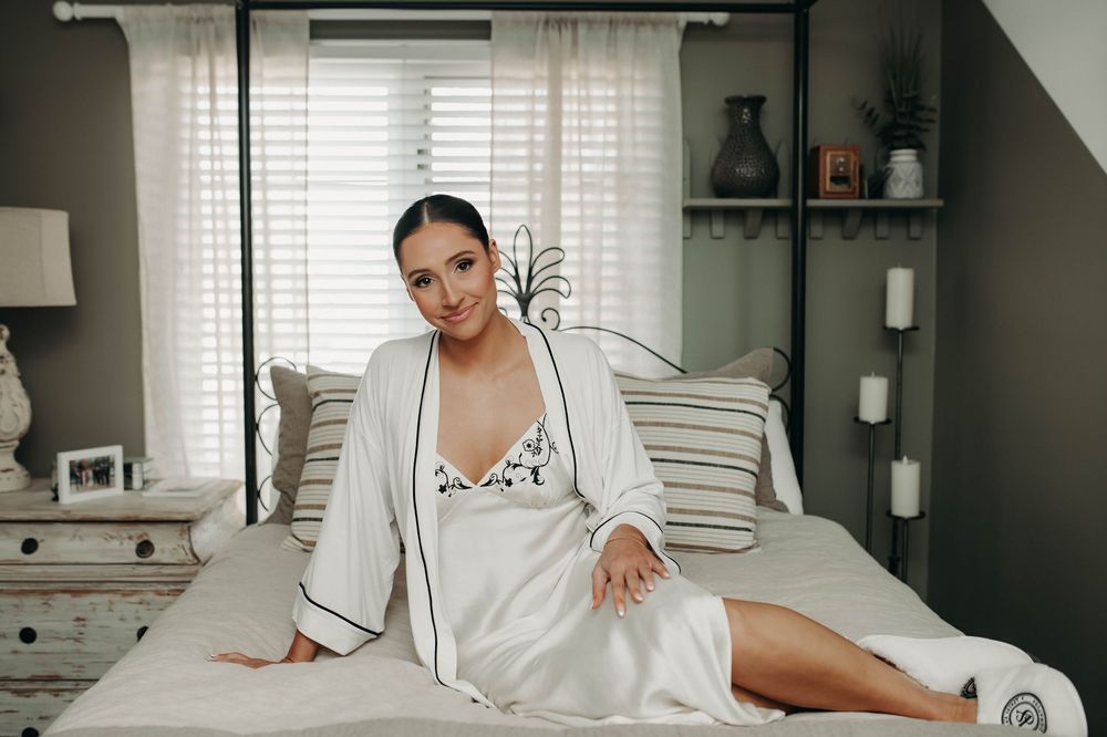 Bride in white satin robe and floral slip sitting gracefully on bed during wedding morning prep.
