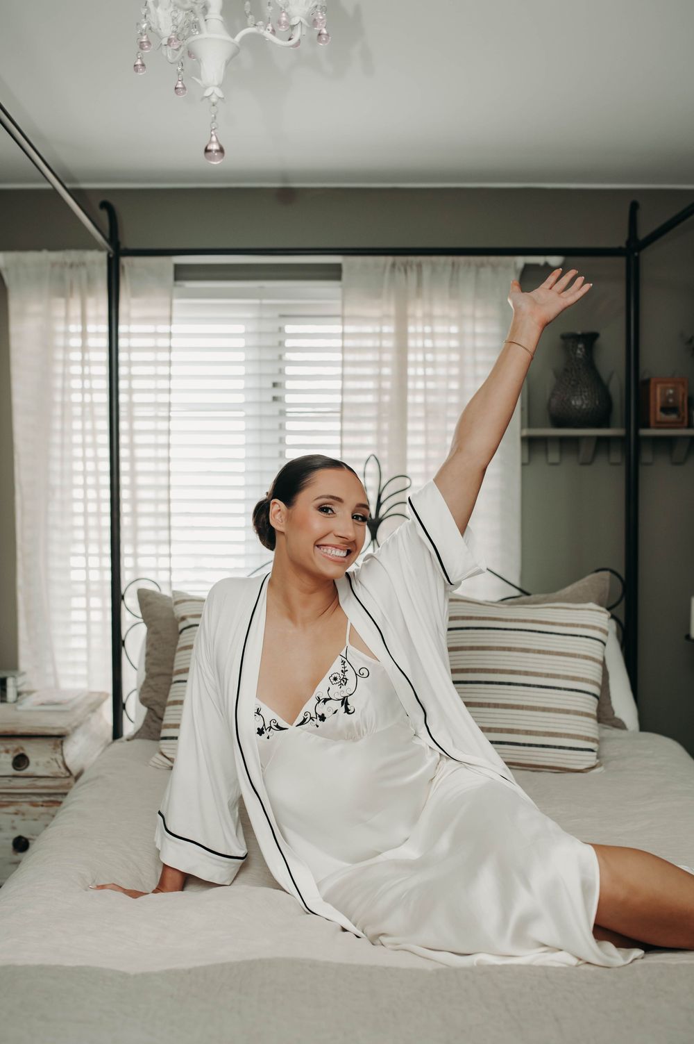 Joyful bride in white satin robe sitting on bed with arm raised in excitement on wedding day.