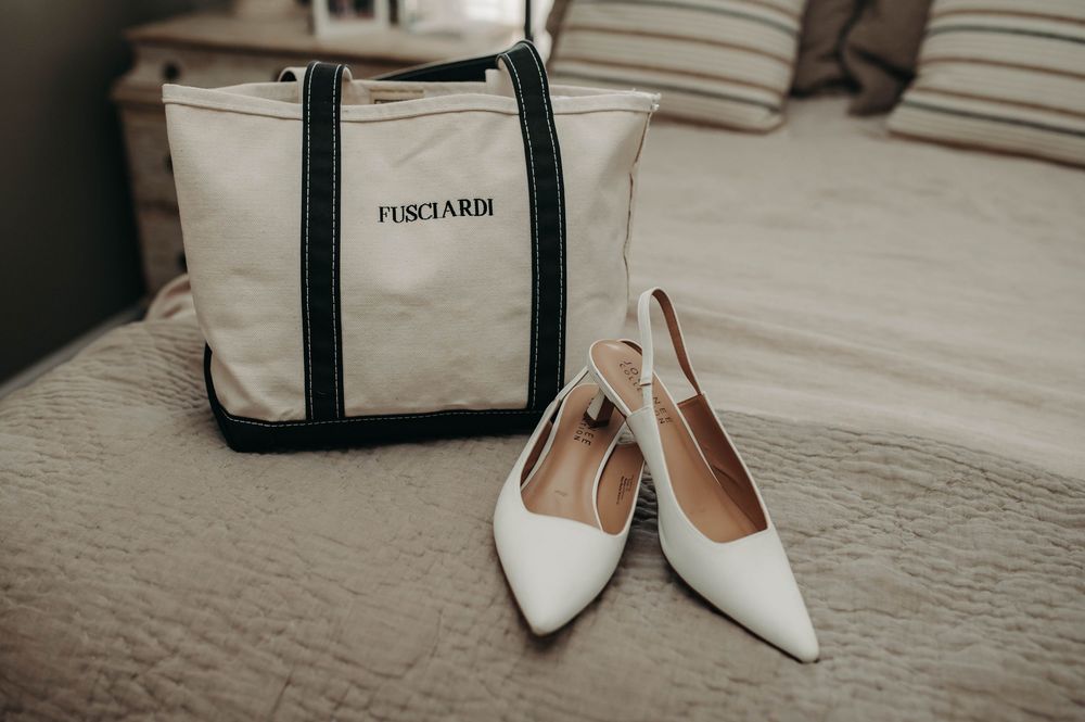 White pointed-toe bridal heels beside a Fusciardi canvas tote bag on a bed.