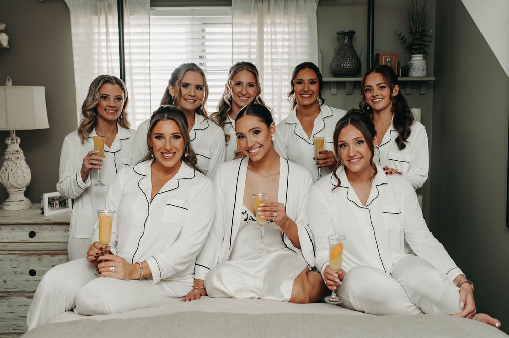 Bride and seven bridesmaids in matching white pajamas celebrating with champagne on wedding morning.