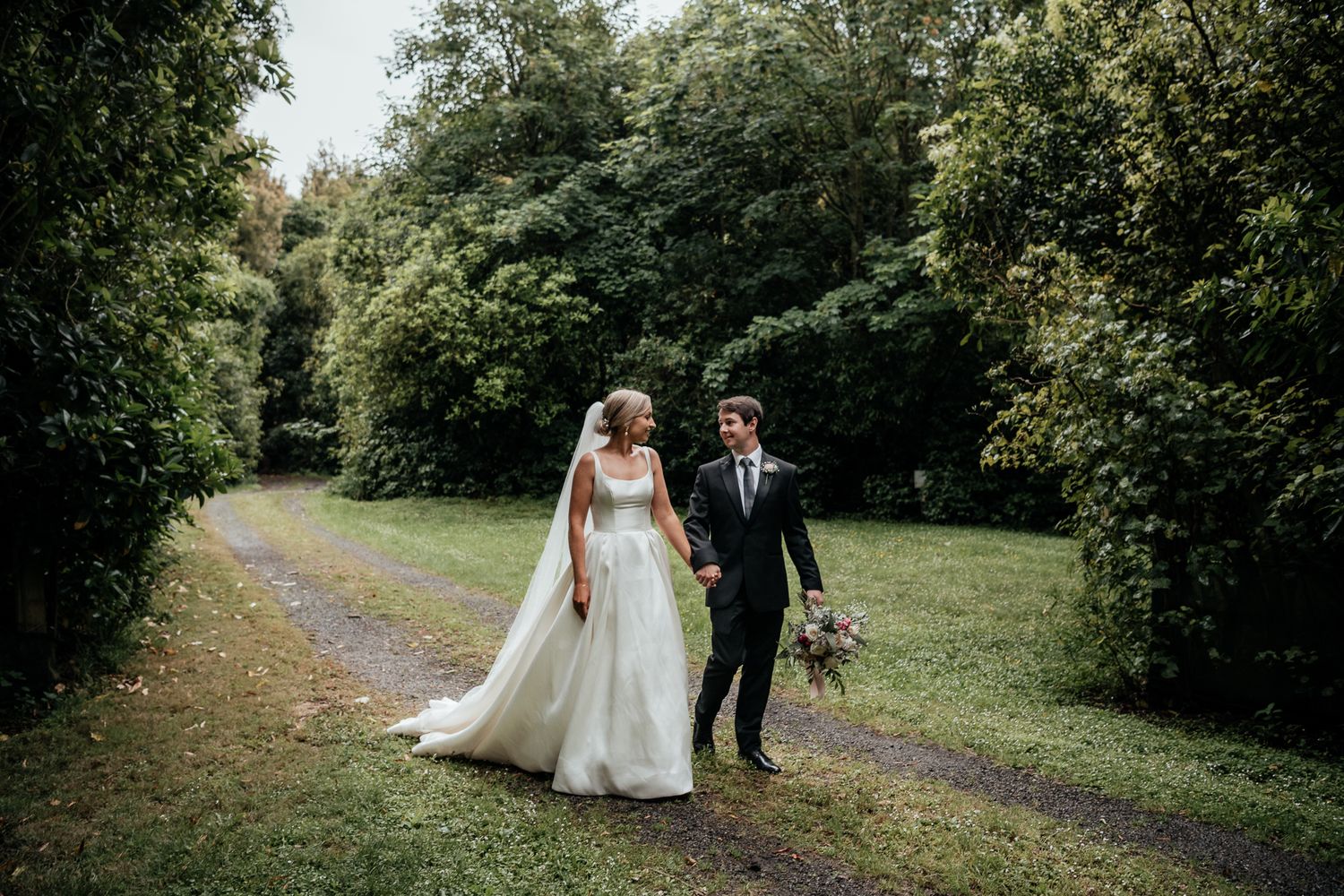 A wedding couple walk together on a tree-lined path in a lush green setting at Rose and Smith  captured by August and Vine