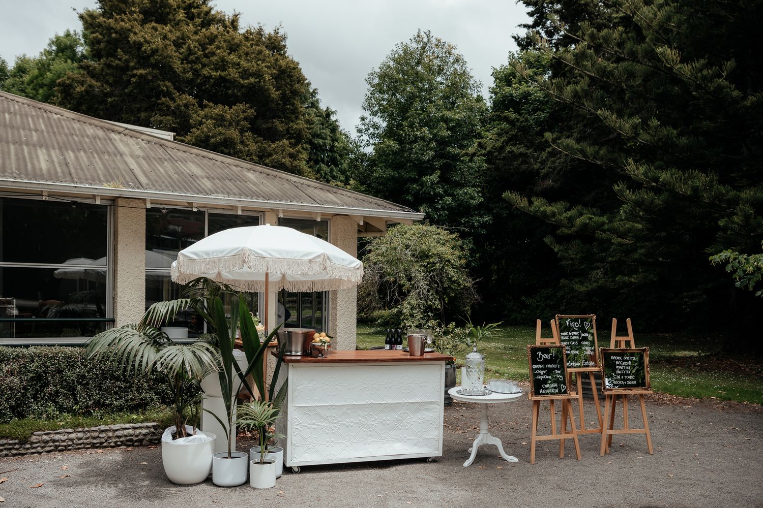 Outdoor wedding venue with cocktail bar, tropical plants and umbrella in a garden setting at Rose and Smith captured by August and Vine