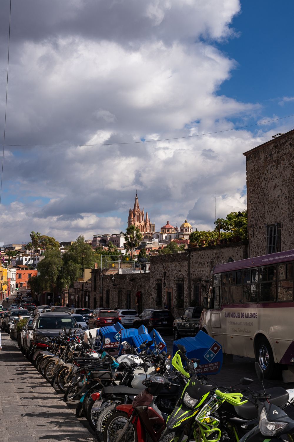 Row of motorcycles parked near stone buildings with a Gothic church spire visible on the hilltop in San Miguel de Allende.